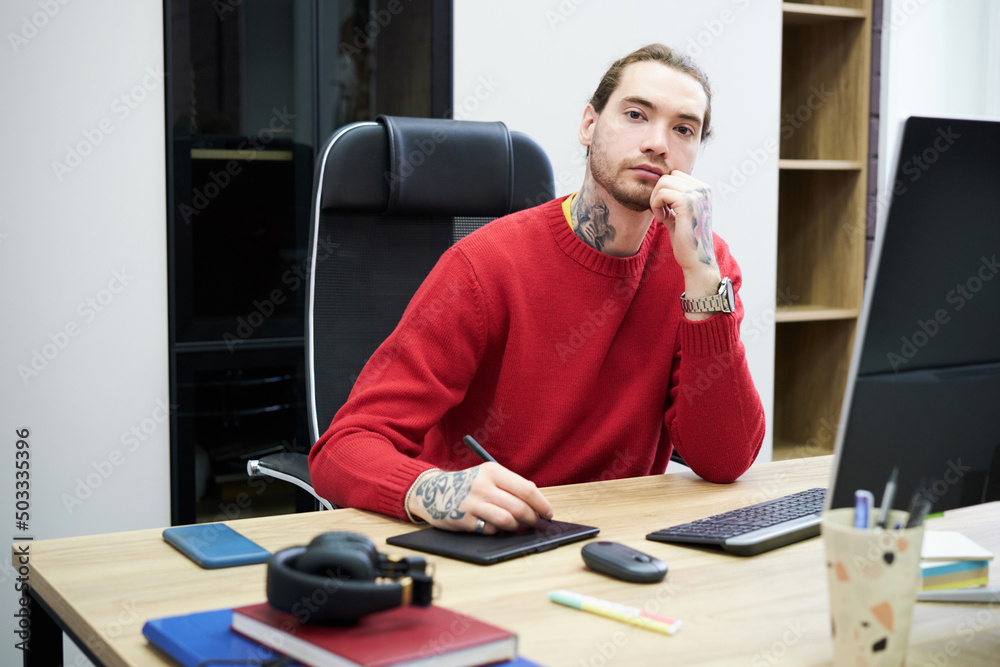 Poster Portrait of young graphic designer looking at camera while sitting at table with computer and working on graphic tablet