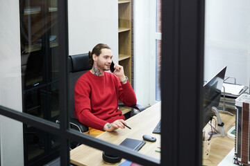 Smiling manager sitting at table at office in front of computer monitor and having business conversation on mobile phone