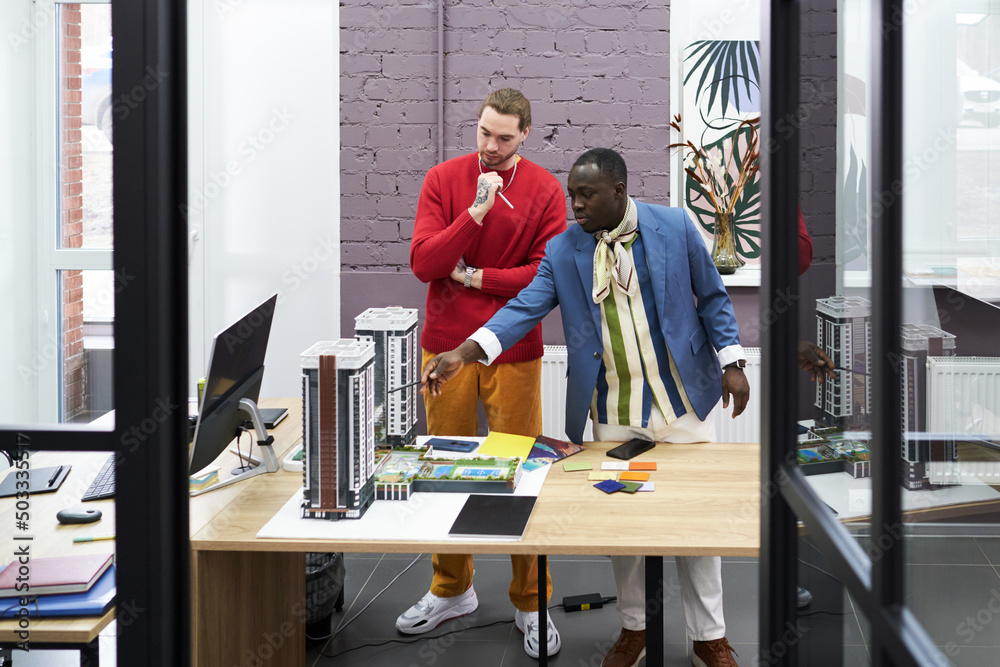 Poster african young architect pointing at mock-up of buildings on table and talking to his colleague
