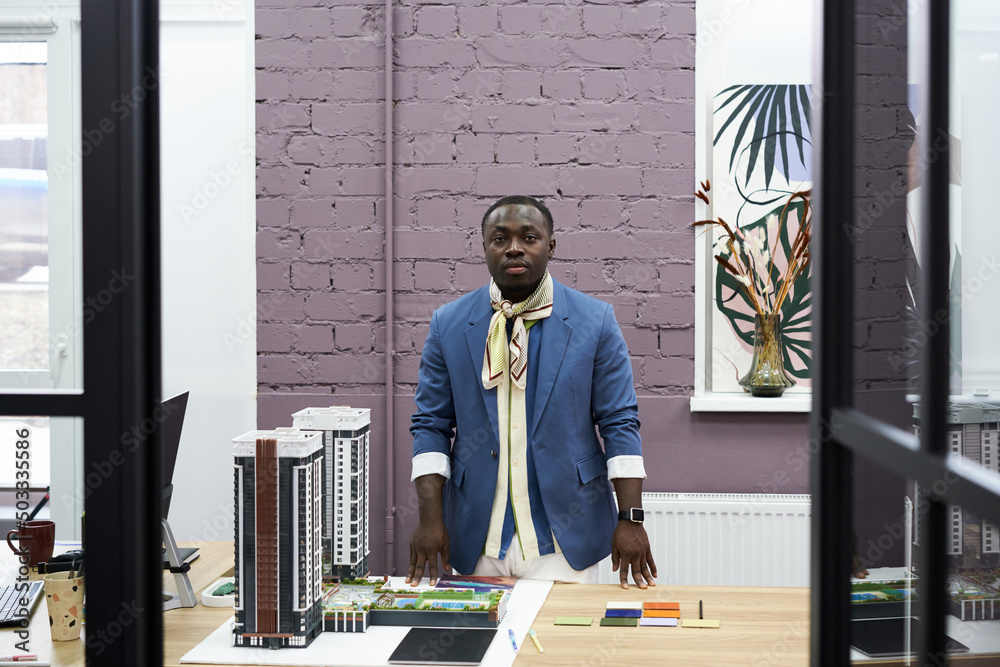 Poster Portrait of African professional architect looking at camera while standing at table with his project of new buildings