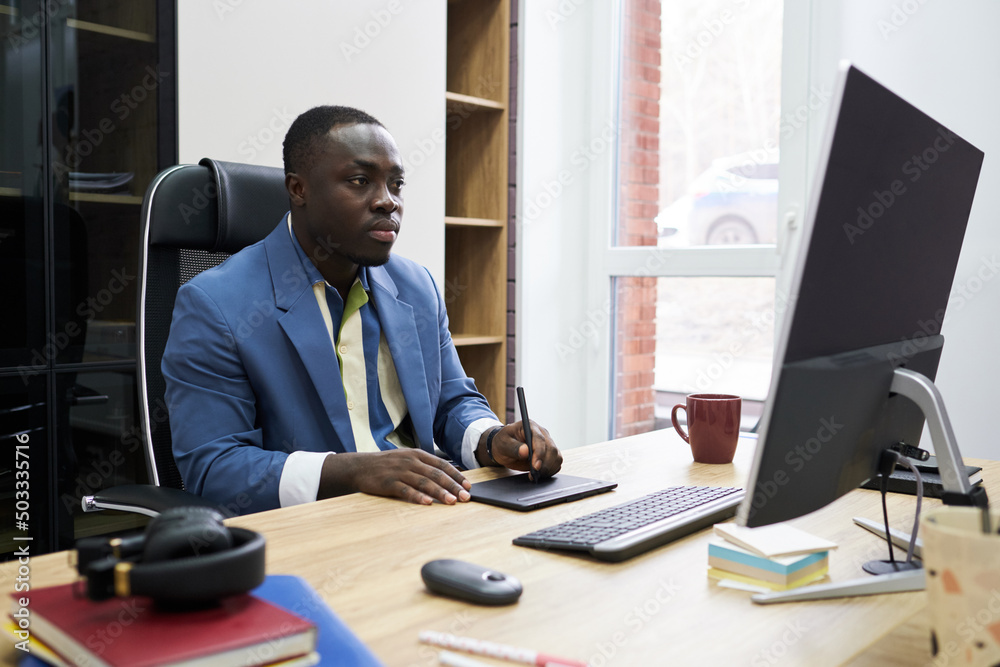 Poster Serious African designer looking at computer monitor while working on graphic tablet at table at office