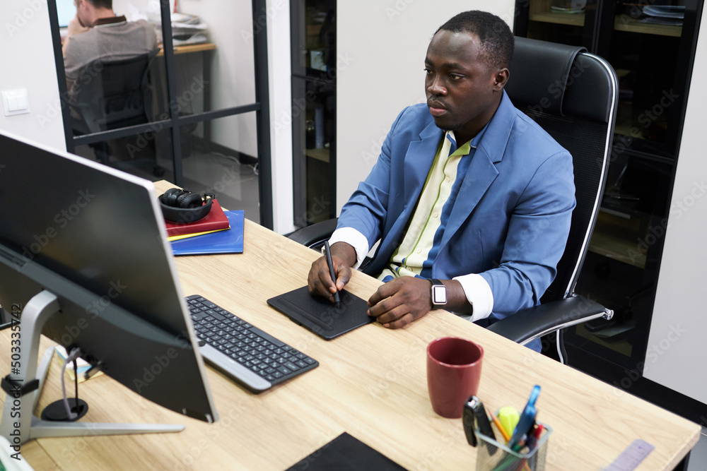 Poster Serious African designer sitting at table at office in front of computer monitor and using graphic tablet for editing