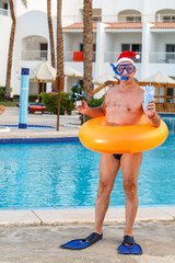 A man in flippers and a mask with a swim ring and a santa hat by the pool on Christmas holidays with a Christmas tree and a cocktail in his hands.