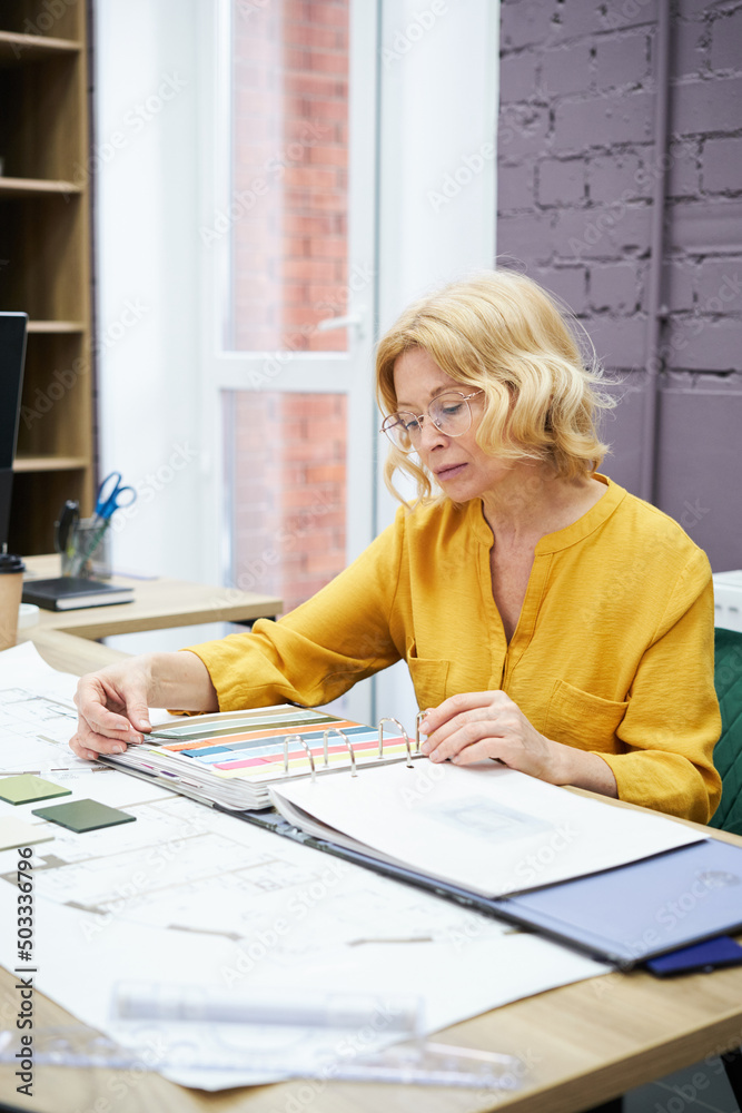 Poster serious female designer in eyeglasses sitting at table and examining colours in palette for future p