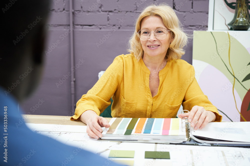 Poster mature interior designer sitting at table with palette and discussing colour with her client during 