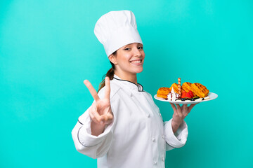 Young caucasian chef woman holding waffles isolated on blue background smiling and showing victory sign