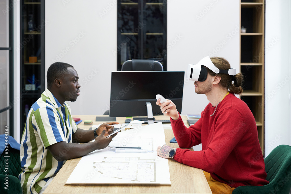 Poster business colleagues sitting at table opposite each othe and using virtual reality goggles at their w