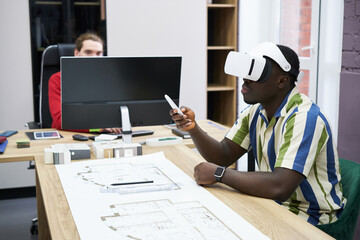 African young architect using virtual reality headset to look at construction project while sitting at table with blueprint