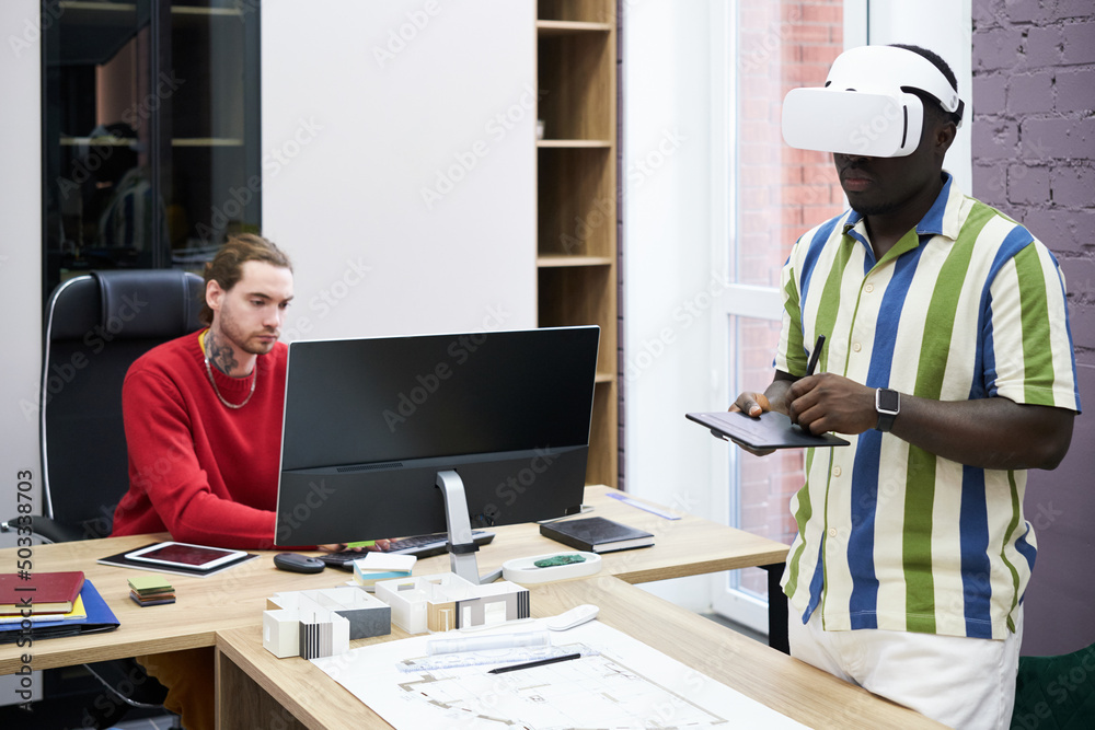 Poster Graphic designer using VR headset to draw blueprint with tablet with his colleague using computer at table
