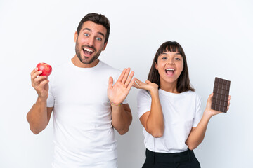 Young couple holding chocolate and apple isolated on white background with surprise and shocked facial expression