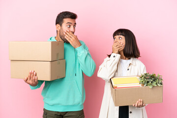 young beauty couple moving in new home among boxes isolated on pink background covering mouth with hands for saying something inappropriate