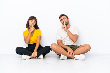 Fototapeta na wymiar Young couple sitting on the floor isolated on white background showing a sign of closing mouth and silence gesture
