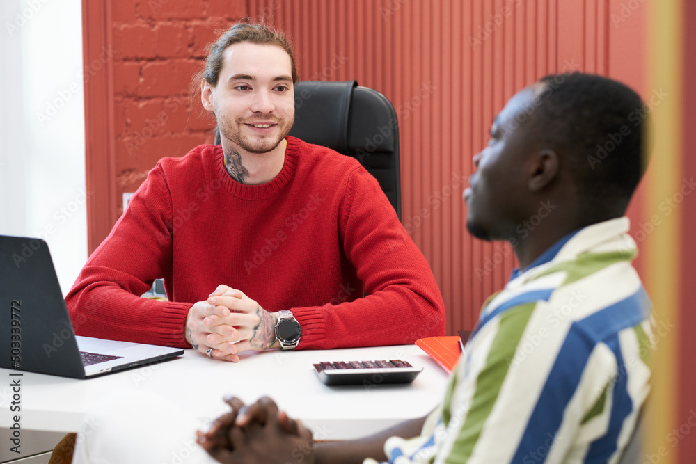 Poster young smiling businessman sitting at table with laptop and talking to client during their meeting at