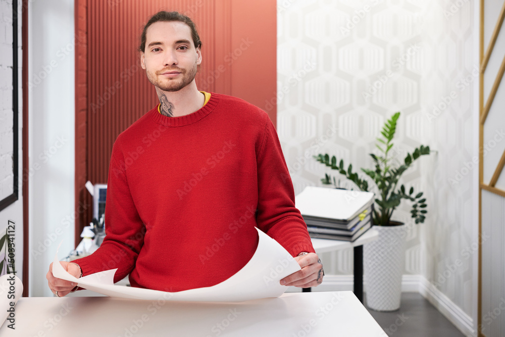 Poster portrait of young designer looking at camera while working with sketches at office