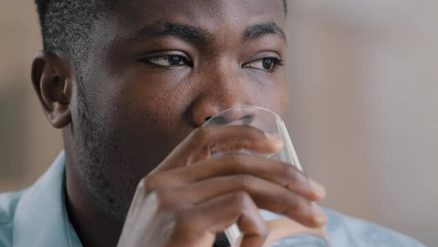 Close up headshot african millennial man drink pure fresh cold water glass healthy liquid suffer from hot make daily habit hydrate thirst american thoughtful businessman guy lost in thoughts look away