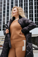 young blonde woman in trendy black quilted coat jacket oversized city walk advertising banner model plus size