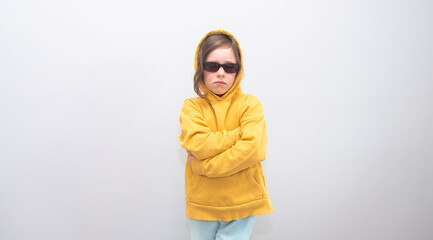 Child, Caucasian 6 years old, in a yellow sweatshirt with a hood and sunglasses on a gray background. Emotions on her face and crossed her arms on the gruli.