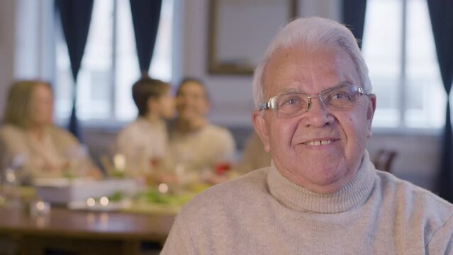 Senior white-haired man in glasses looking at camera and smiling while having nice time at family party. Blurred family image in background. Family reunion, bonding, retirement concept