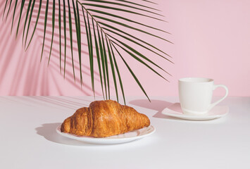 Tropical palm leaf, fresh croissant and cup of coffee on pastel pink background. Minimal food composition. Breakfast concept.