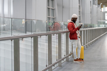Thoughtful African American traveler man using smartphone at airport terminal, travelling with luggage bag. Passenger guy waiting her flight, checks time of flight online, standing in railway station