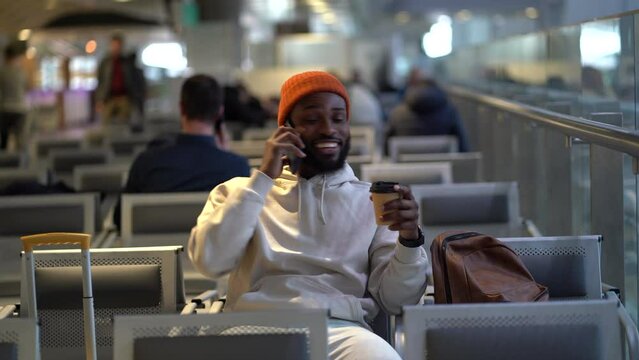 Smiling young African American traveler man drinking coffee while waiting for flying at airport terminal. Handsome black hipster guy talking on cellphone, drinking tea and looking away. 