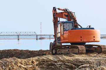 An orange excavator with a hydraulic bucket works in the morning at a construction site to strengthen the river bank. Road bridge and power line in blur in the background.