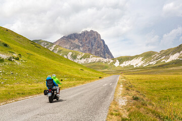 Two people riding a motorcycle on a mountain road. A couple of tourists on motorcycles on a road in the Apennines in Abruzzo, Italy. Campo Imperatore plateau with the Gran Sasso peak in the background
