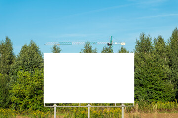 Blank white billboard for advertisement in front of trees. There is also tower crane in the background. Idea of city development.