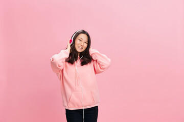 Obraz na płótnie Canvas Enjoying Asian student young lady in pink hoodie sweatshirt with cute headphones smile touch head posing isolated on over pink studio background. Good offer. Sound streaming platform ad concept
