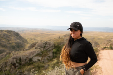 Portrait of Latin woman dressed in black with a cap having fun during the day of trekking in the mountain forest - looking at the horizon