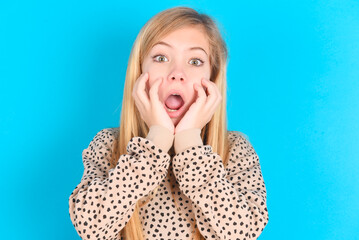 Stupefied little caucasian kid girl wearing animal print sweater over blue background expresses...