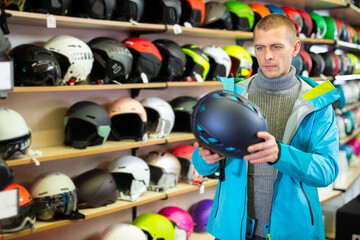 American man buyer in blue ski jacket chooses helmet in a sports equipment store