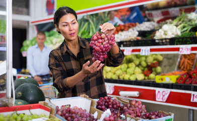 Interested asian woman visiting greengrocery store, choosing ripe sweet grapes from cardboard...