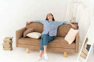 Obraz premium Calm young woman resting on a soft comfortable sofa, sitting enjoying a carefree peaceful morning, relaxing on the sofa