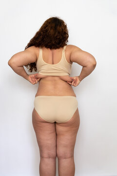 Back view of overweight middle-aged woman with long curly hair wearing beige underwear, standing, squeezing, stretching skin of back to show subcutaneous fat on white background. Weight loss, obesity.