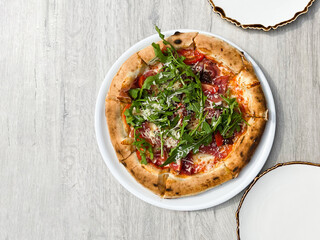 Top view of pizza with prosciutto, arugula, tomatoes, and parmesan on a white plate on the table. Cooked. Cuisine. Tasty.  Round. Green. Healthy. Italian. Delicious. Plate. White. Ham. White Plate