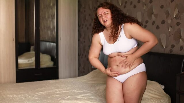 Curly overweight suffering woman in white underwear stand near bed in bedroom and with pain on face press on stomach. Fat burning treatment of cellulite body. Weight loss process, diet and exercise.
