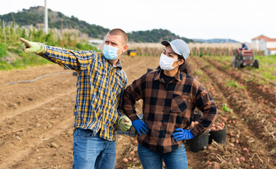 European man and Asian woman farmers in face masks standing on potato field. Man pointing with finger.