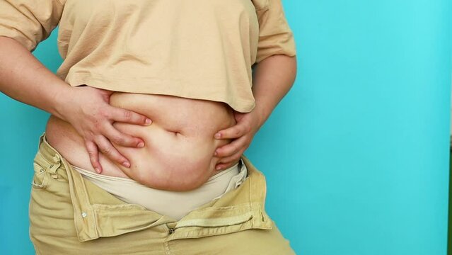 Unrecognizable plus-size woman wearing beige T-shirt, unzipped jeans, squeezing, crumpling excess flabby stomach on blue background. Body positive, massage, obesity, weight loss, liposuction, diet.