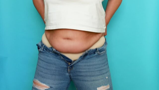 Unrecognizable fat plump overweight woman wearing beige underwear, white short T-shirt, putting on blue jeans, squeezing belly on blue background. Body positive, obesity, weight loss, liposuction.