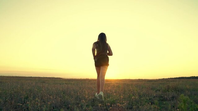 Jogger girl breathes fresh air on field. Free young woman runs in summer park at sunset. Running after sun. Training jogging. Healthy beautiful girl is engaged in fitness, jogging in country in sun.