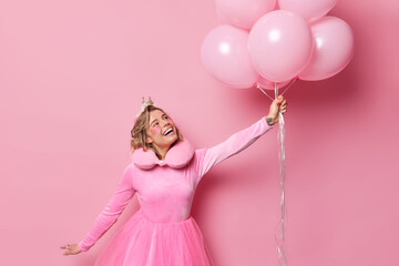 Obraz na płótnie Canvas Positive young woman prepares for party undergoes beauty procedures wears festive dress holds bunch of balloons floating in air isolated over pink background. People and special occasion concept