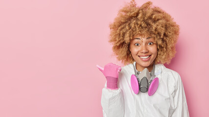 Positive woman in white protective costume uses gas mask points away on blank space against pink background tells about radiation hazard protects herself from environmental pollution. Look there