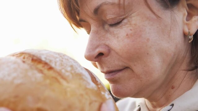 Mature woman baker smelling round loaf of fresh bread at sunset. Slow motion.