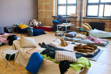 Large group of sleeping places with blankets, pillows, toys, sacks and other essentials prepared for refugees in spacious room - obrazy, fototapety, plakaty