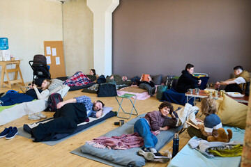 Several refugees having rest on their sleeping places on wooden floor in spacious room while young woman talking to her son with book