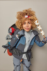 People active rest leisure and lifestyle concept. Serious curly female camper poses with roasted marshmallow spends free time on summer camping carries backpack isolated over grey background