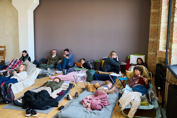 Group of refugees using mobile gadgets, talking and sleeping while having rest on couchettes and mattresses in spacious room