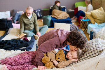 Young woman in casualwear kissing her cute little son sleeping with teddybear while bending over...