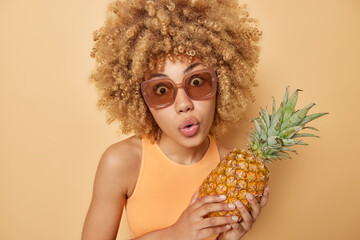 Portrait of surprised woman keeps lips rounded looks with amazement holds pineapple wears trendy sunglasses and casual t shirt isolated over beige background. Female model with exotic fruit.